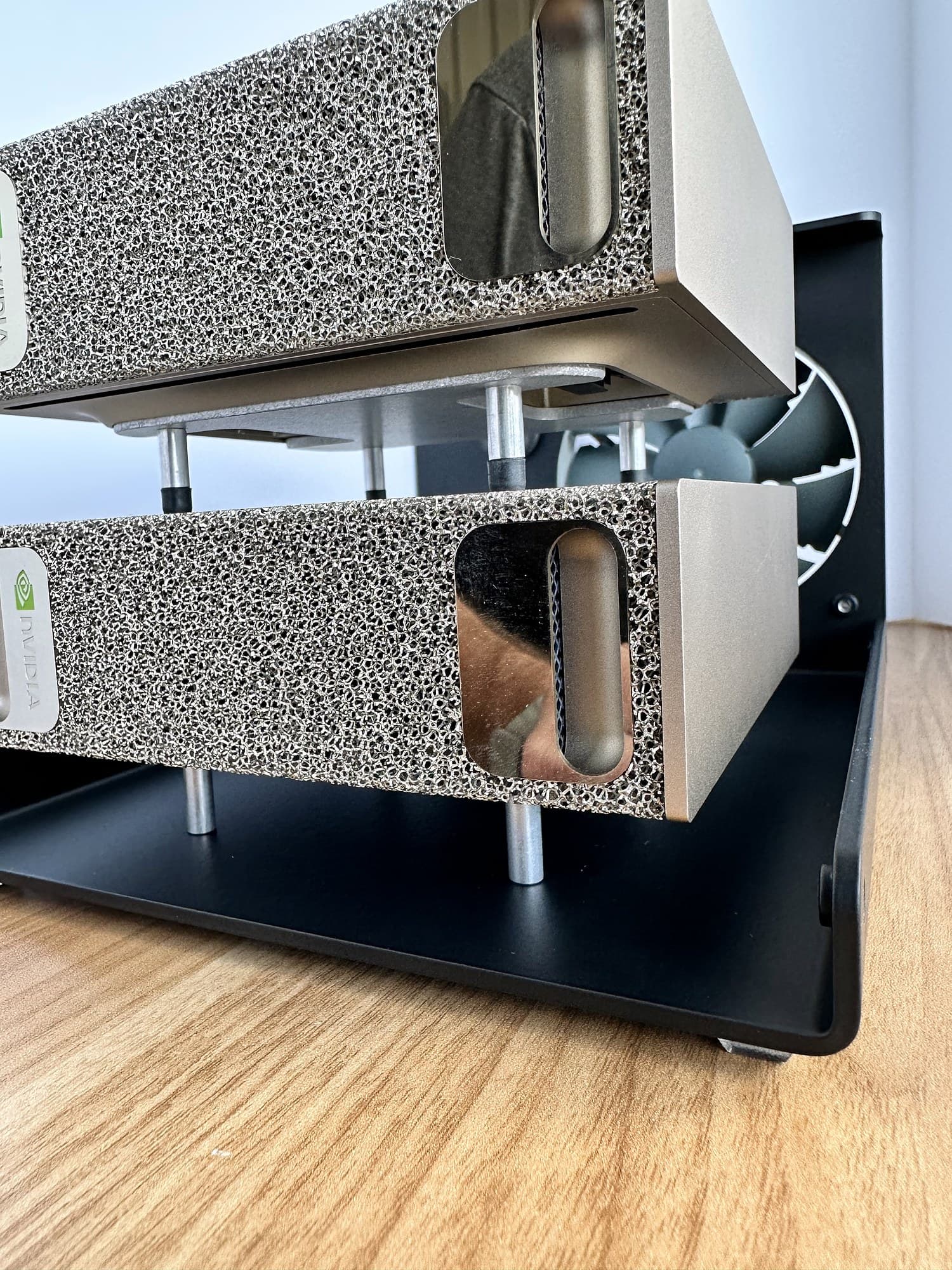 Close-up of two NVIDIA DGX Spark units stacked on the standoffs in the black Stackcooler base, with the rear fan behind, on a wood desk.