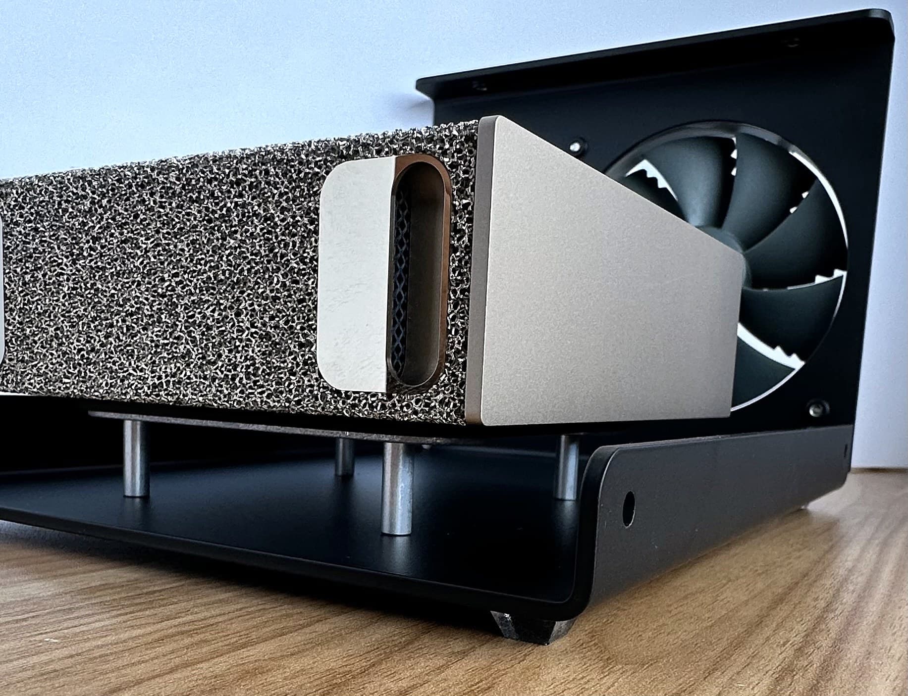 Low-angle view of the bottom DGX Spark resting on the H-bracket standoffs inside the black Stackcooler base, with the rear fan behind it, on a wood desk.