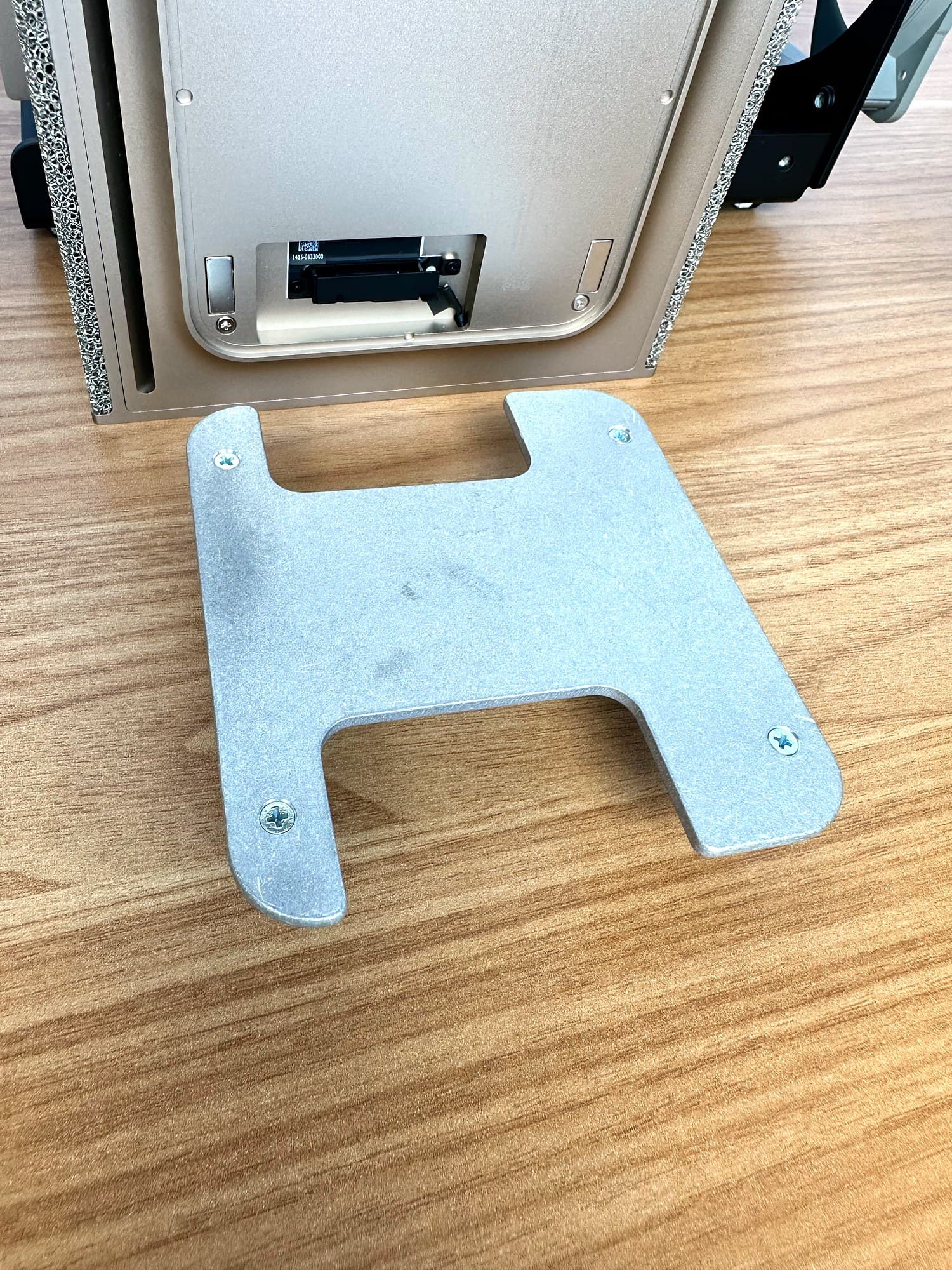 Aluminum H-bracket with its screws showing, lying on a wood desk beside the underside of the top DGX Spark with its pad removed.